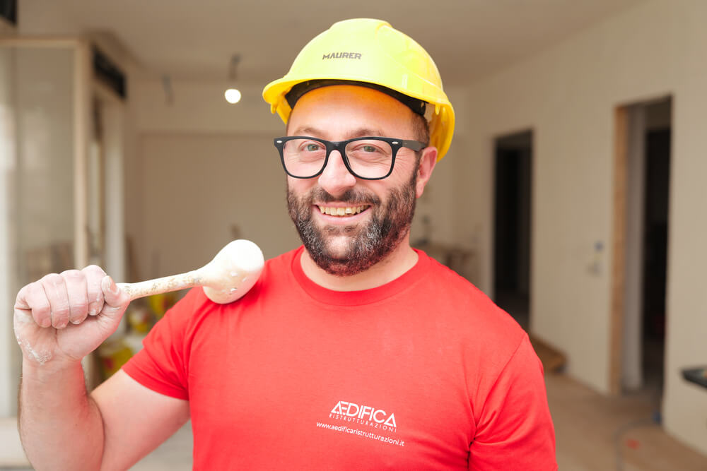 Operaio edile sorridente, casco giallo, maglietta rossa.